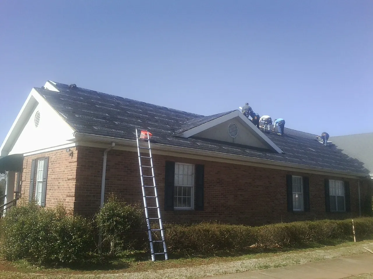 Crew tearing shingles off a brick ranch roof with a ladder set — Metal Roof Repair work by our crew in Bainbridge